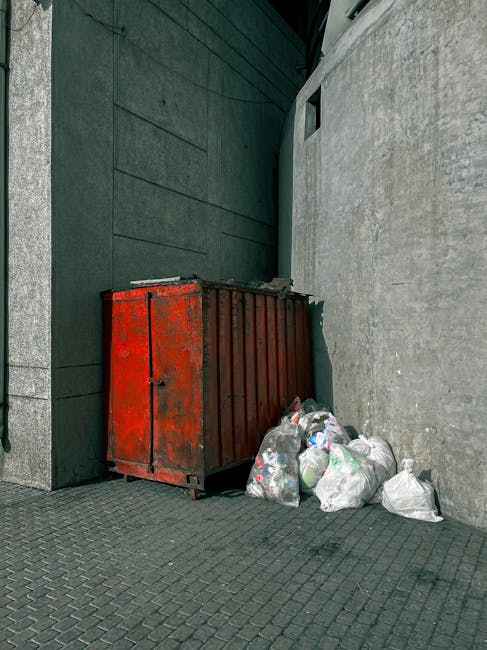 A white waste collection truck is parked on an urban street in front of a brick building with multiple windows, some of which have white frames and glass panes. The truck is equipped with a rear-loadi
